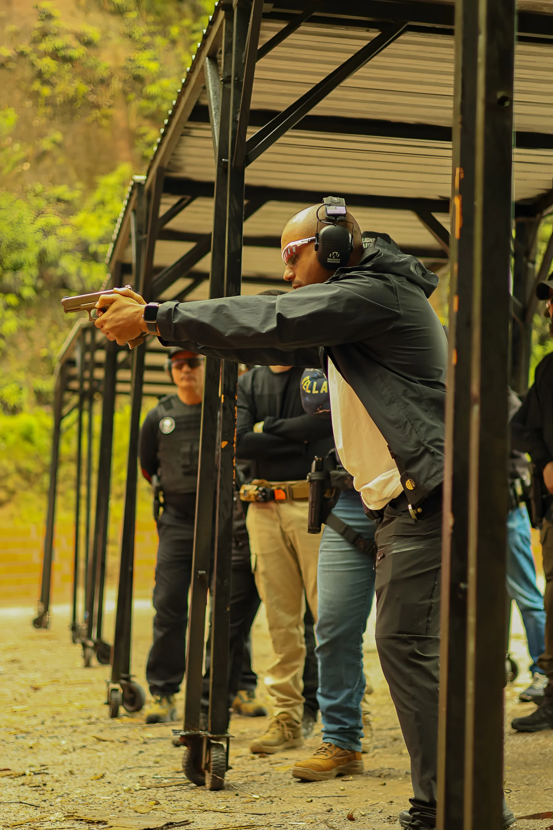 Instrutor Neres atirando com a pistola na baia coberta do estande, com alunos observando ao fundo